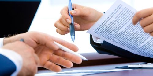 A close-up of two people's hands over a desk during a meeting. One person holds a blue pen and a printed document, while the other gestures with a white pen toward papers on the table.